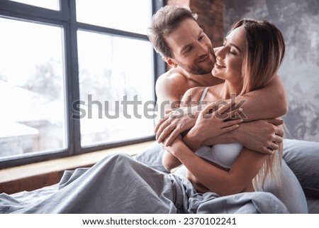 Beautiful couple is smiling and hugging while spending time together in bed at home. Man is kissing his girlfriend