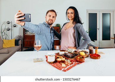 Beautiful Couple Smiling With A Positive Attitude For A Selfie At Home In Front Of A Table With Tapas Food. Millennials, Technology, Relationship, Blogging, And Foodie Concept