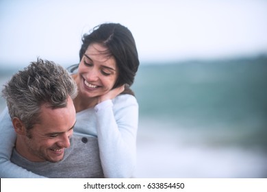 A Beautiful Couple In Their Forties Walks On The Beach, The Man Wears The Woman On His Back, They Wear Sweaters And Jeans