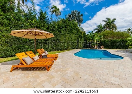 Beautiful courtyard with pool area, sun loungers with umbrella, covered patio with outdoor furniture, privet wall, palms, short grass, blue sky