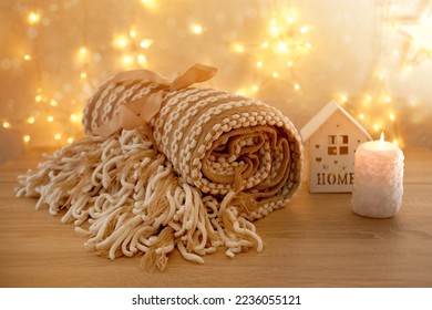 beautiful cozy creamy plaid with fringe lies rolled up on light table against festive background with bokeh of garlands, wooden house with candle, concept of sweet house, warm cute gift for holiday - Shutterstock ID 2236055121