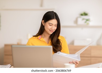 Beautiful Creative Asian Young Woman Using Laptop Computer And Open Document To Reference Her Work.Young Freelancer Woman Wearing Yellow Shirt Sitting At Desk Working Online From Home.Small Business