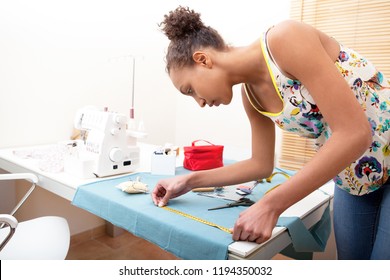 Beautiful creative young fashion design college student using measuring tape, making clothes, interior. Skills and trades, learning concentration. Black female seemstress working at home, indoors.
