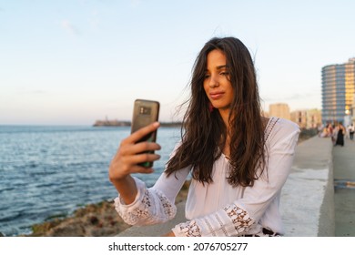 Beautiful Cuban Woman Portrait On The Malecon Using Smartphone. Havana, Cuba. Real Life Style Image At Sunset.