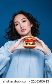 A Beautiful Curly Asian Girl With Bright Makeup Wearing A Blue Hoodie Holding A Big Homemade Burger With Two Hands Over A Navy Blue Background