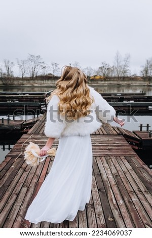 A beautiful curly blonde bride in a white fur coat walks along a wooden pier outdoors. Wedding photography, portrait.