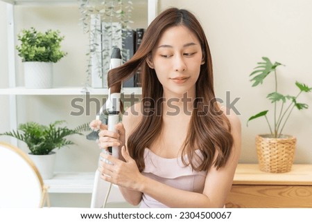 Beautiful curly brunette, attractive asian young woman, girl smiling using curler, curling iron long hair with ceramic curler straightener at home. Hairstyle and Hairdressing. Beauty and hair care.