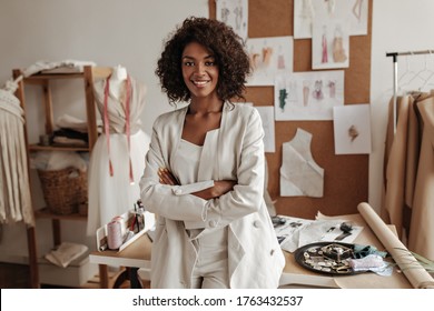 Beautiful curly brunette dark-skinned fashion designer poses in office, leans on table. Young lady in white suit crosses arms and smiles.