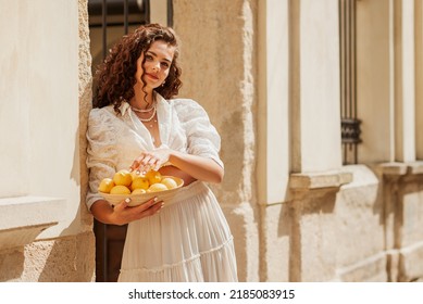 Beautiful curly brunette woman wearing trendy summer white linen shirt, skirt, holding bowl with lemons, posing in sunny street of European city. Copy, empty space for text