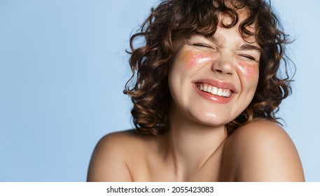 Beautiful curly girl smiling with white teeth, glitter on clean natural facial skin, laughing happy, standing over blue background