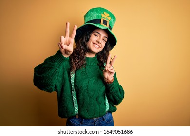 Beautiful curly hair woman wearing green hat with clover celebrating saint patricks day smiling looking to the camera showing fingers doing victory sign. Number two.