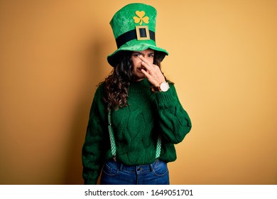 Beautiful Curly Hair Woman Wearing Green Hat With Clover Celebrating Saint Patricks Day Smelling Something Stinky And Disgusting, Intolerable Smell, Holding Breath With Fingers On Nose. Bad Smell