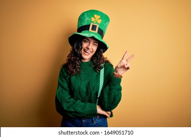 Beautiful curly hair woman wearing green hat with clover celebrating saint patricks day smiling with happy face winking at the camera doing victory sign. Number two.