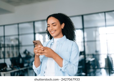 Beautiful curly haired multiracial young business woman in stylish elegant shirt, team lead, business coach, secretary, standing in modern office, using smartphone, messaging with clients, smiling - Powered by Shutterstock