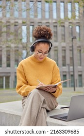 Schöne, geschliffene Frau hört Webinar Noten in Notizbuch arbeitet an Projektargen aus gestricktem gelbem Pullover und Hosen verwendet Laptop-Computer-Posen gegen Gebäude in der Stadt Arbeiten Fernbedienung