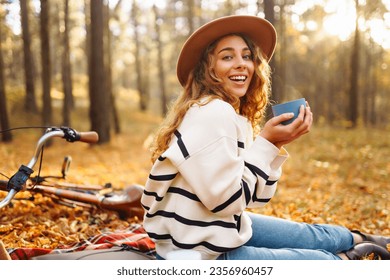 Beautiful curly woman in a hat sits on a red mat with a thermos in the autumn forest. Female enjoys nature at mini picnic. Concept of relaxation, nature. - Powered by Shutterstock