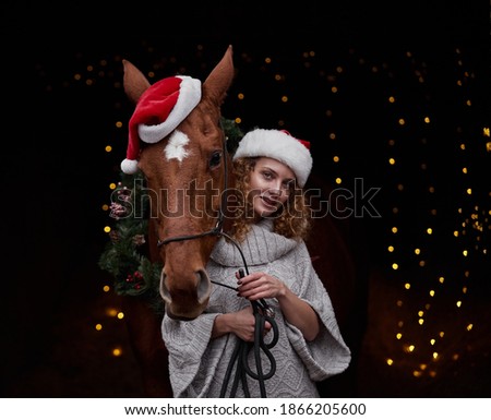 Beautiful curly-haired girl in a Santa hat with a red horse in a hat on a black background in New Year's decorations