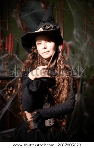 Beautiful curly-haired sorceress in a black hat makes a spell with a magical book and a wand in a dark old castle covered in cobweb. Art of witchcraft. Magic Halloween spells. 