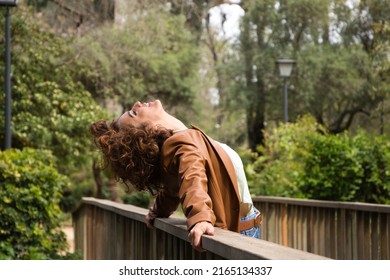 Beautiful, curly-haired woman leaning on the railing of a wooden bridge with her head tilted back. The woman is dressed in modern clothes. Travel and holiday concept, rest and relaxation. - Shutterstock ID 2165134337