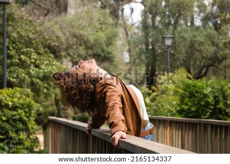 Beautiful, curly-haired woman leaning on the railing of a wooden bridge with her head tilted back. The woman is dressed in modern clothes. Travel and holiday concept, rest and relaxation.