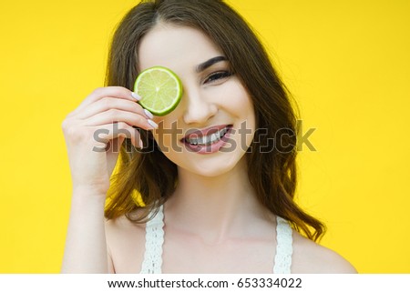 Beautiful cute brunette girl with a lime slice near the face isolated on a yellow background