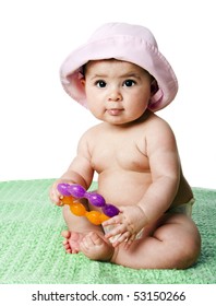 Beautiful Cute Caucasian Hispanic Baby Girl Sitting On Green Blanket Holding An Orange With Purple Teething Ring Toy, Wearing Pink Hat, Isolated.