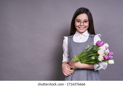 Beautiful Cute Emotional Girl Long-haired Brunette In Glasses, Holds Multi-colored Tulips In Her Hands And Looks At The Camera, Lavish Gift To The Left Of Copy Space,very Joyful, On A Gray Background.