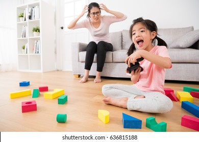 beautiful cute excited daughter playing video games by controller with many messy toy sitting on wooden floor with angry woman mother looking her in living room on background sofa.