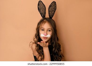 A beautiful cute girl model dressed up as an Easter bunny sends an air kiss  A headdress and black lace ears   painted face child and hare's mustache
