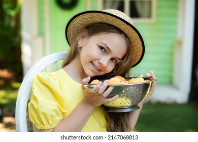 Beautiful Cute Happy Kid Girl With Colander Full Of Apricots Is Gathering Harvest. Closeup Portrait Of Happy Little Girl With Long Hair And Straw Hat, Looking At Camera. Smiling Brunette Child Model. 