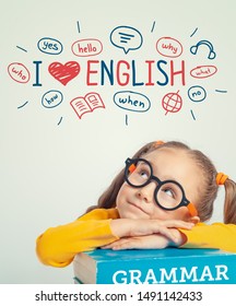 Beautiful Cute Little Girl Leaning On English Grammar Book And Looking At The Illustrations And Words In English Above Her Head. I Love English Concept.