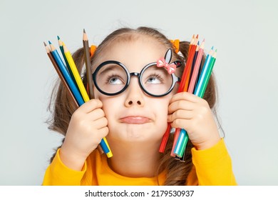 Beautiful cute little preschooler girl with glasses holding colorful pencils and pouting her lips. Sulky child with pencils. Imagination and creativity at school concept. 