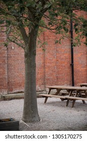 A beautiful dark brown rustic weathered wooden hand made pub table and bench, surrounded by trees and a red brick wall.  - Shutterstock ID 1305170275