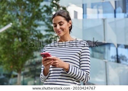 Beautiful dark haired woman holds smartphone texting on modern device wears casual striped jumper walks in street during sunny day visits foreign country uses high speed interntet buys tickets online