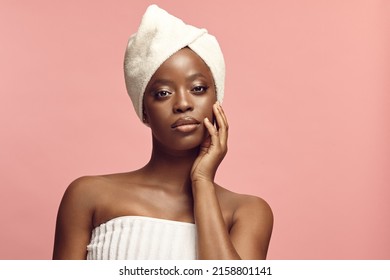Beautiful dark skin woman wearing in body and head towel standing on a pink background touching self face.