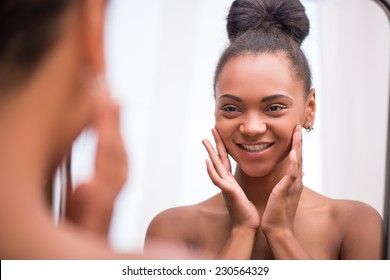 Beautiful  dark skinned girl in white towel bringing face cream looking at mirror admiring  herself  touching cheeks smiling  isolated on white background