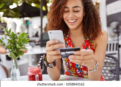 Beautiful dark skinned young female with cheerful expression, holds smart phone and credit card, banks online or makes shopping while sits against cafe interior. Payment and leisure concept.