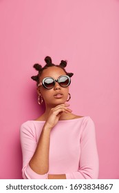 Beautiful dark skinned young woman keeps hand under chin, wears trendy summer sunglasses and stylish jumper, makes fashion photography, models in studio against pink background. Vogue, style