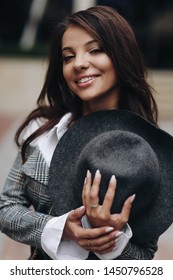 Beautiful dark-haired girl in gray jacet hold a hat on her chest on a spring day. Vertical outdoor close up portrait of cute woman looking at camera and smiling.