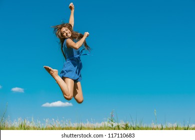 Beautiful dark-haired happy young woman jumping high in air, against background of summer blue sky.