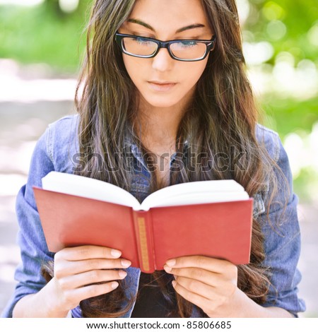 Beautiful dark-haired serious girl in jeans jacket and glasses reads red book against summer green park.