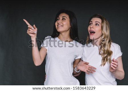 Beautiful dark-haired smiling Asian woman standing with surprised blonde friend and pointing away with amazement in studio on dark background