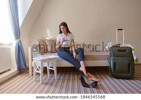 A beautiful dark-haired woman is sitting on a sofa in a hotel room. She took off her shoes after a day of work and continues to work, staying in touch with the office