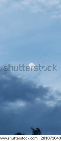 Beautiful daytime view of the moon shining through a blue sky with dramatic dark clouds. Ideal for weather, astronomy, nature, and atmospheric background concepts.