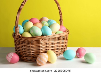 Beautiful decorated Easter eggs in wicker basket on white wooden table against yellow background, closeup
