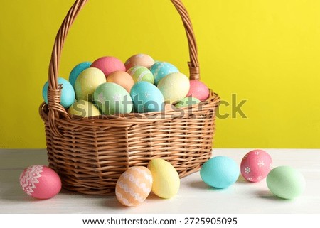 Beautiful decorated Easter eggs in wicker basket on white wooden table against yellow background, closeup