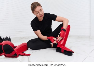 Beautiful determines athletic woman boxer fighter putting on red kickboxing leg pads guard. Sparring shin guards, isolated over white background. Martial art, combat, challenge. Kickboxing concept.