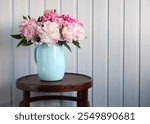 A beautiful display of peonies in shades of pink and white sits in a stylish blue jug atop a wooden table, creating a warm and welcoming ambiance in a cozy indoor setting.