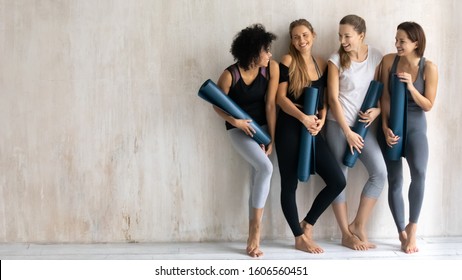 Beautiful Diverse Girls With Yoga Mats In Hands Standing At Grey Fitness Center Wall Background With Copy Space, Young Sporty Women Wearing Sportswear Chatting And Laughing After Training