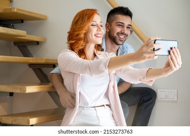 Beautiful Diverse Young Couple Sitting On Stars At Home And Taking A Selfie Photo On Smartphone. Red Haired Woman Holding Smartphone To Take Self Portrait And Embracing With Her Boyfriend